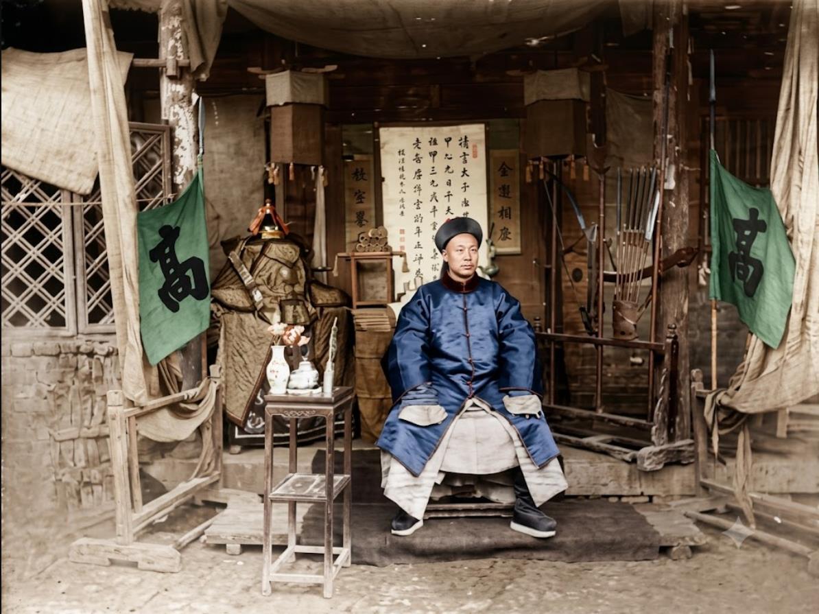 清末，山东邹平，高姓男子端坐堂前，身旁立着醒目的“高”字大旗，身后摆满棉甲、佩刀