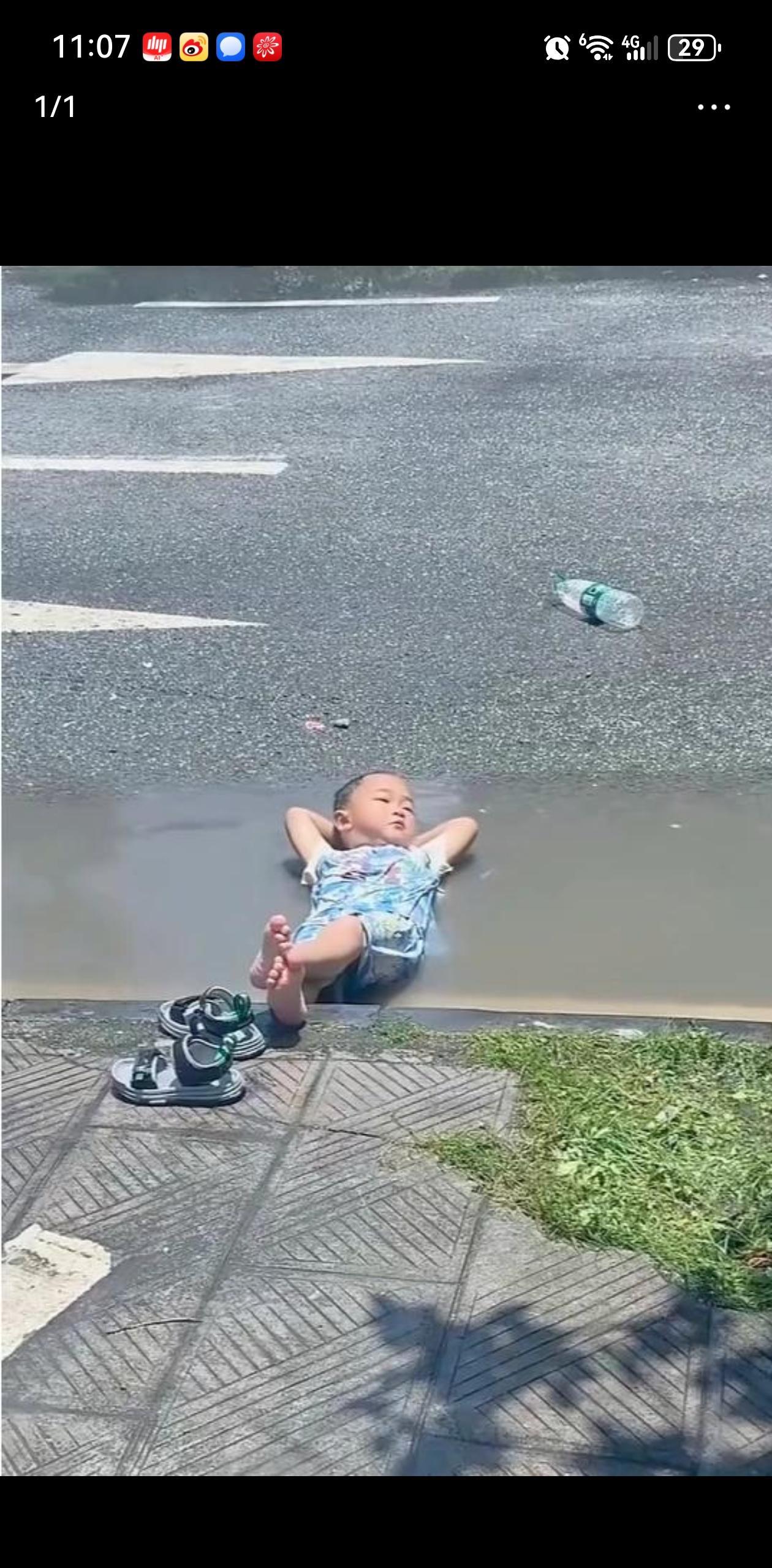 一小男孩头枕双手躺在马路牙子旁边的积水里，二郎腿一翘，悠哉悠哉，简直不要太爽！