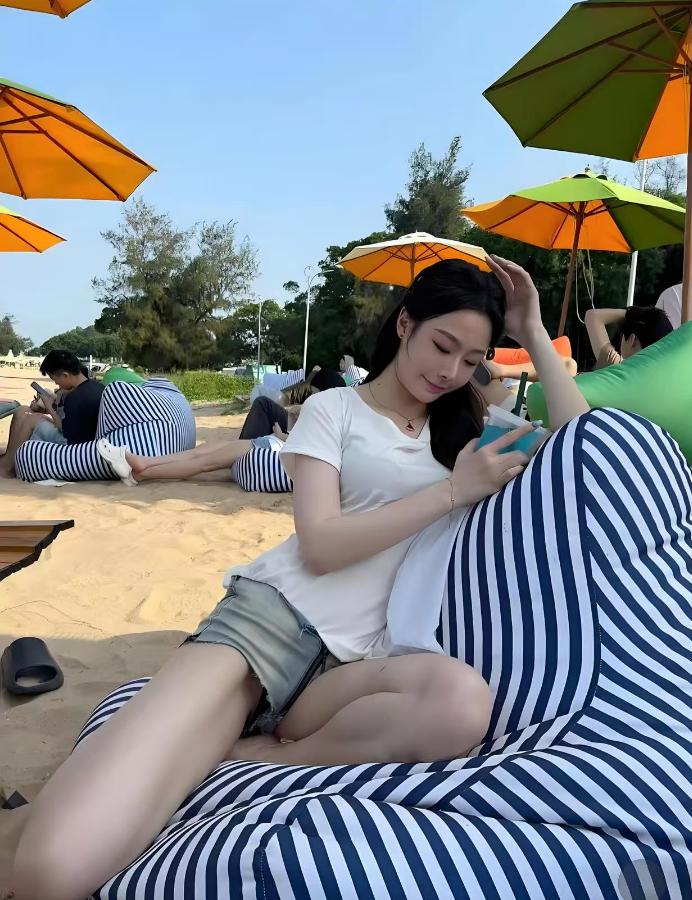 这沙滩上的画面也太惬意了吧——蓝天底下，彩色遮阳伞衬着松软沙粒，她窝在条纹懒