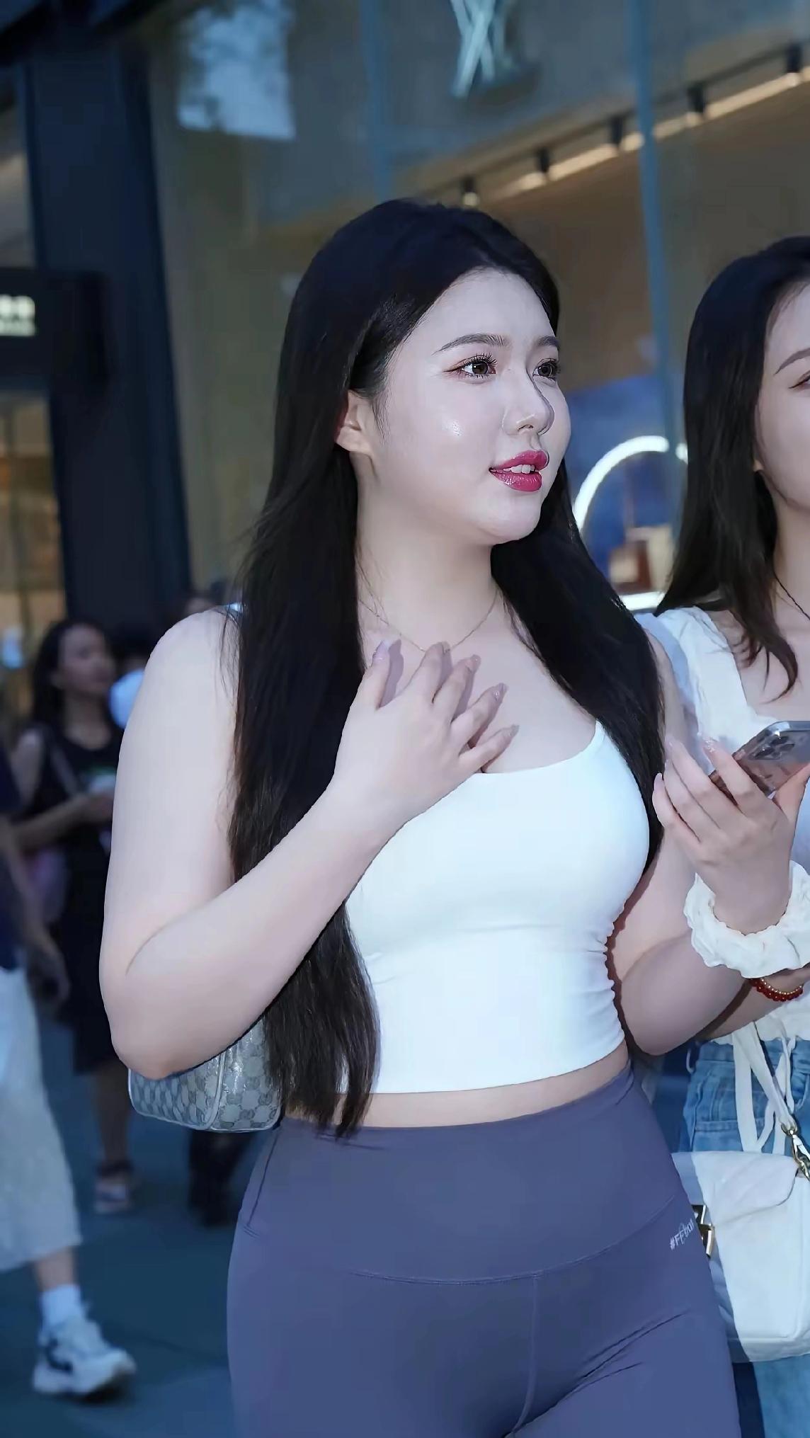 她穿白色短款背心，搭配深灰紧身瑜伽裤，斜挎一个灰白花纹小包，长发垂肩，唇色红润，