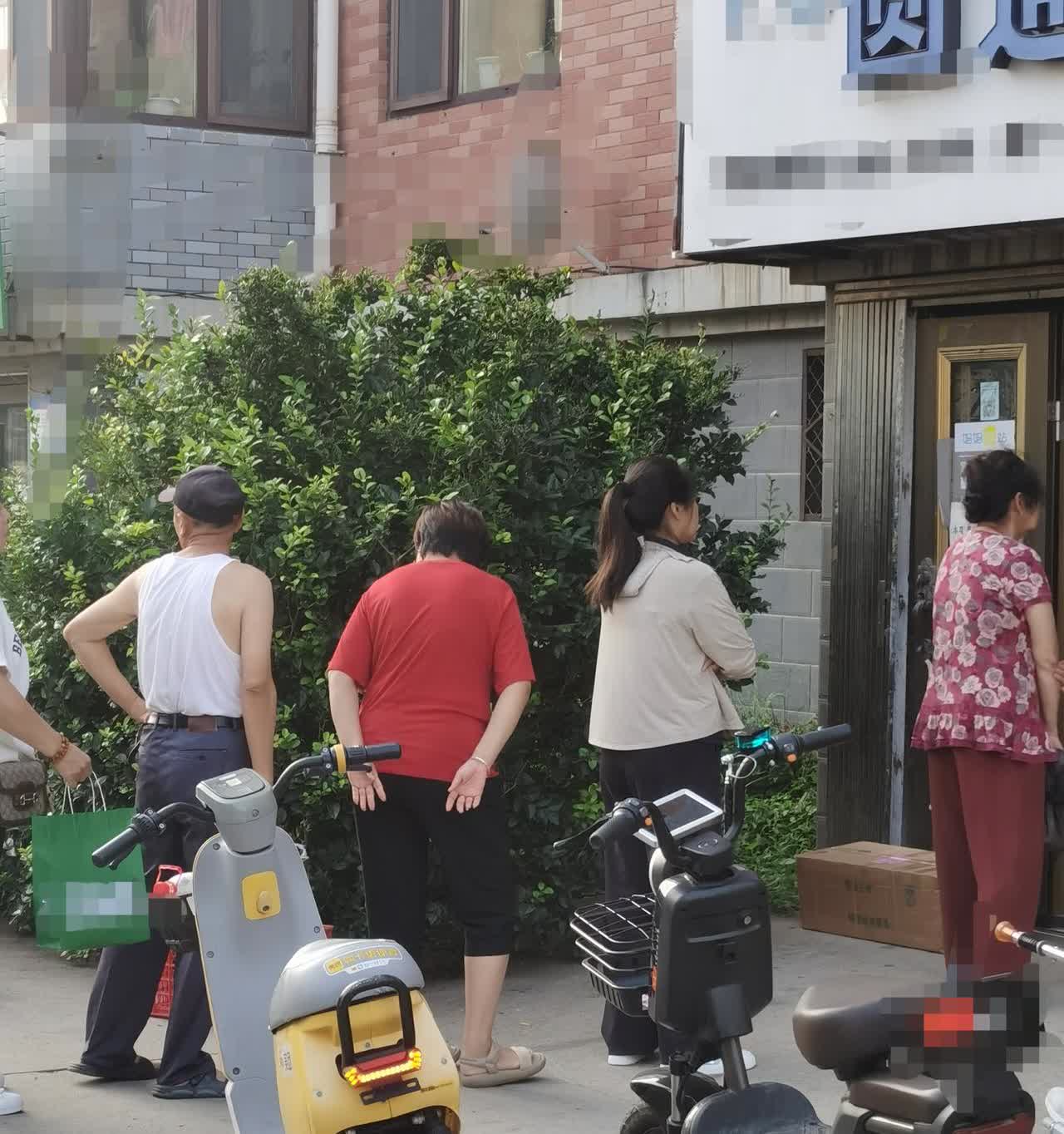 一年轻女子骑着一辆共享电摩车在一驿站外排队取快递的队伍前停了下来，有个刚取完快递