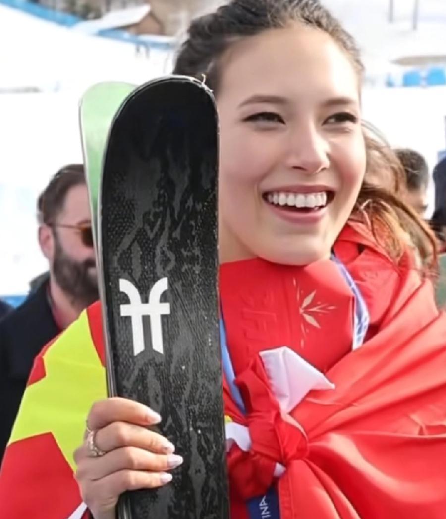 谷爱凌，身披中国国旗五星红旗，参加颁奖后互动，脖子挂着银牌，拿着滑雪板，笑得非常