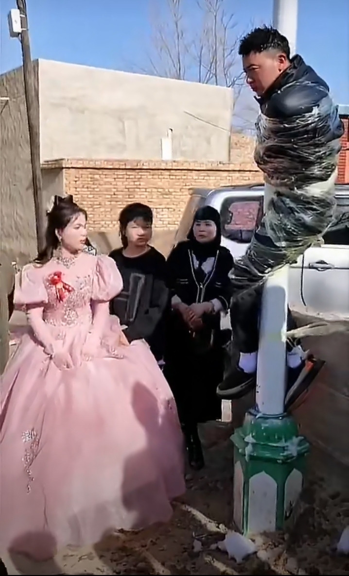 某地婚闹一幕新郎官被人用胶带缠在电灯杆子上，像猴一样挂着，有人拿着皮带之类的东西在身上抽打。旁边的新