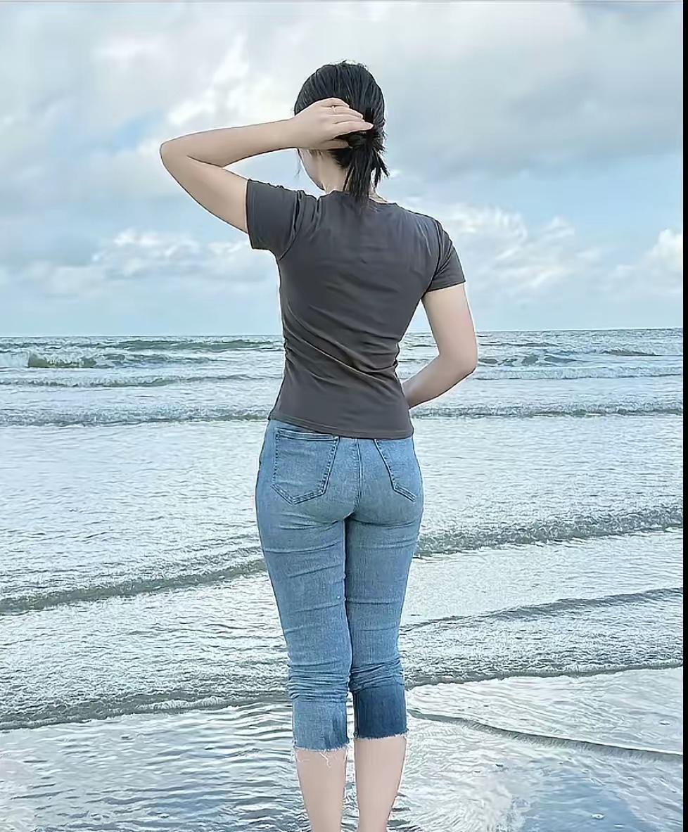 她立于滩涂，身影被海风轻揉。灰衫贴合脊背，勾勒出时光浅淡的轮廓，七分牛仔裤裁剪着