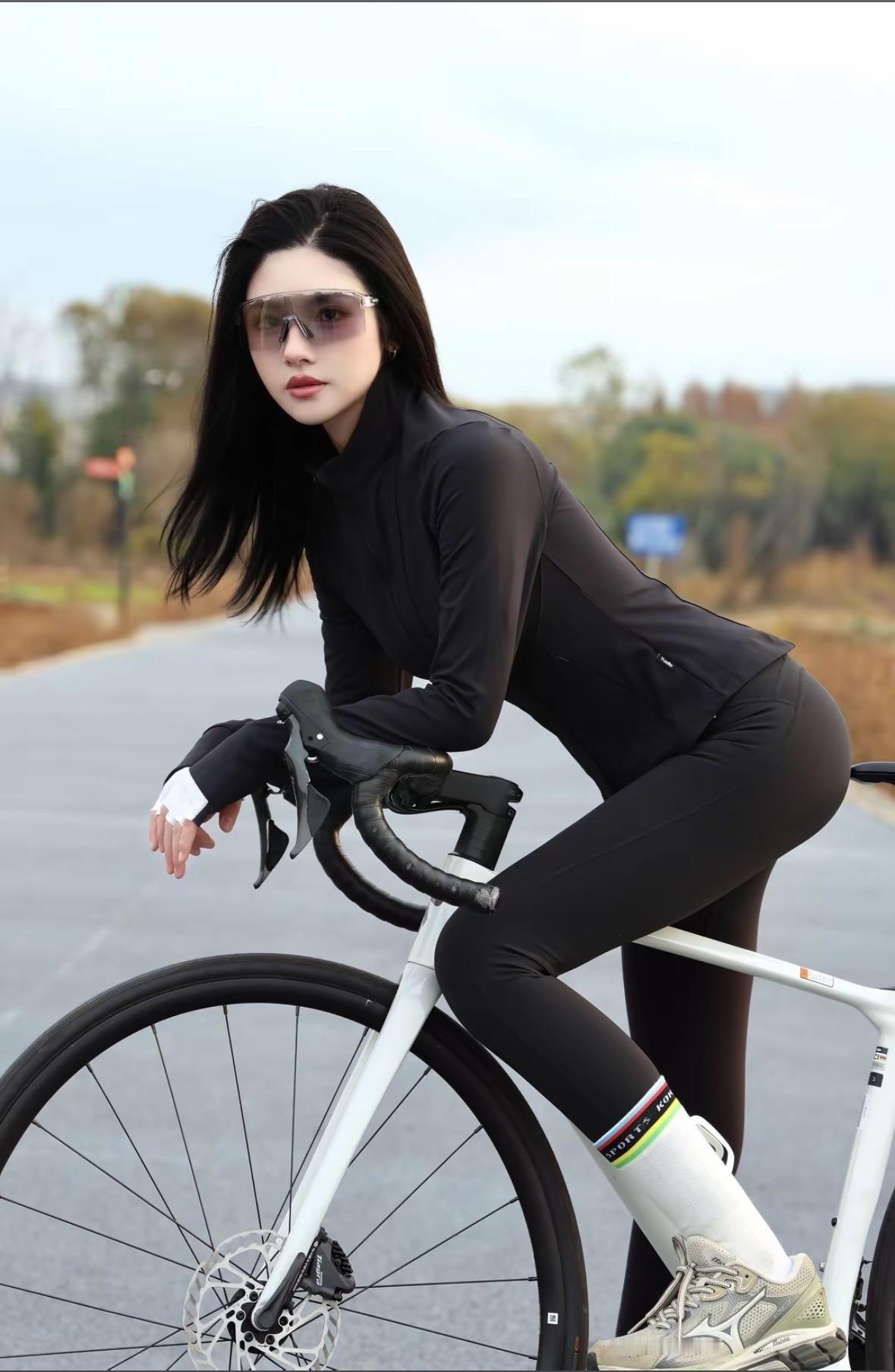 黑发随风飘逸，她身着紧身黑骑行服，佩戴银色风镜，倚靠在白色自行车旁。利落的短发与