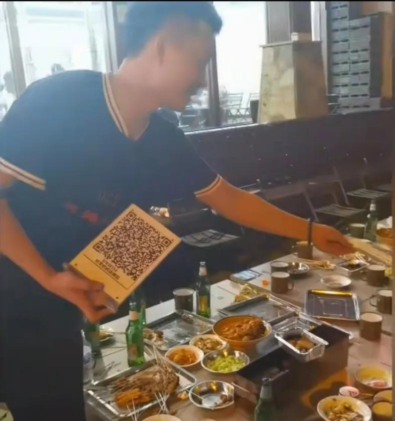 在街头的夜市摊上，老板正拿着收款二维码要坐在摊前的一个小伙子结账，小伙子看着满桌
