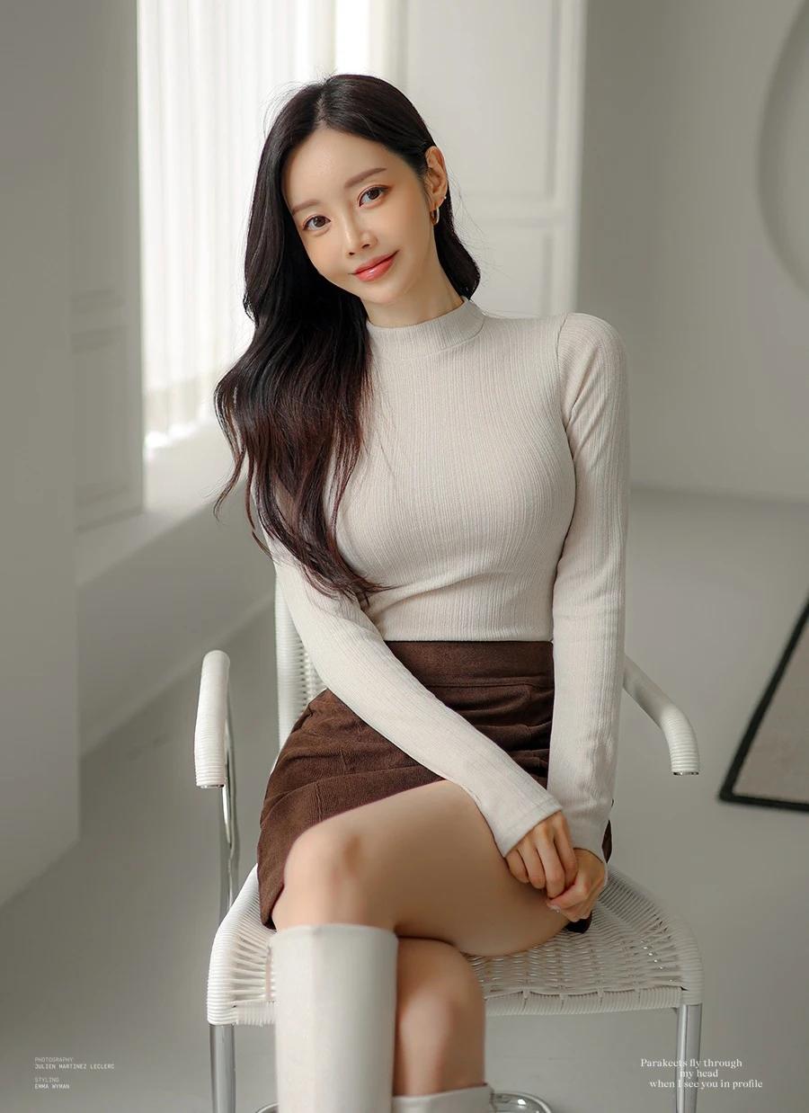 孙允珠-温柔甜美的少女，白色毛衣搭配棕色短裙，坐在白色椅子上，微笑着看向镜头。你