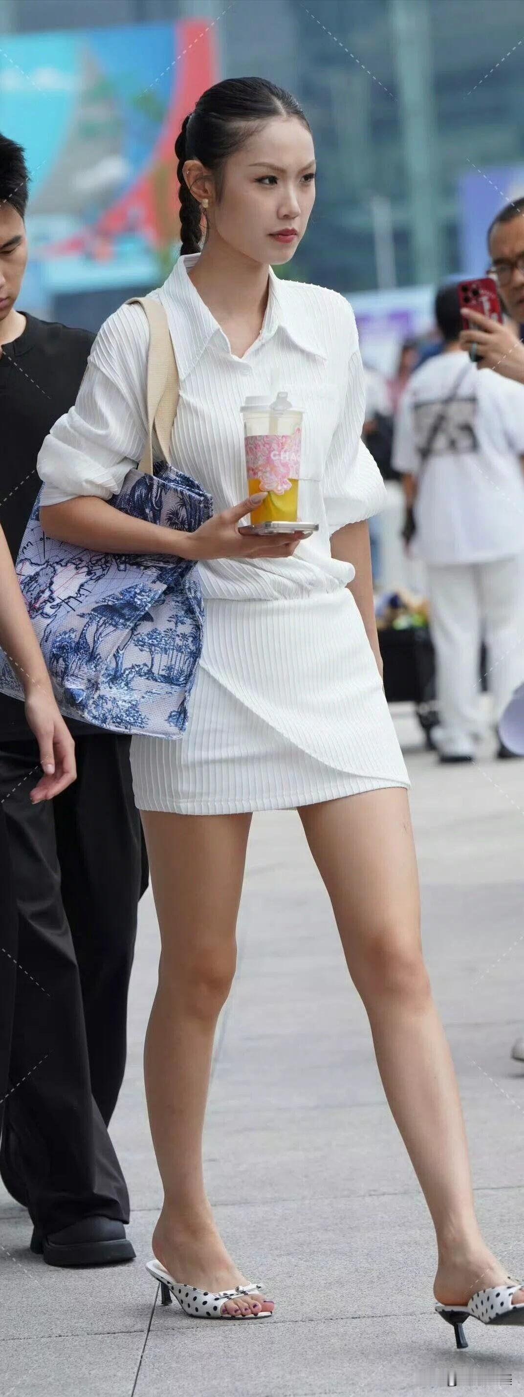 街头偶遇一身白裙的时尚女孩，利落编发搭配波点高跟，手提饮料与印花包，简约又高级，