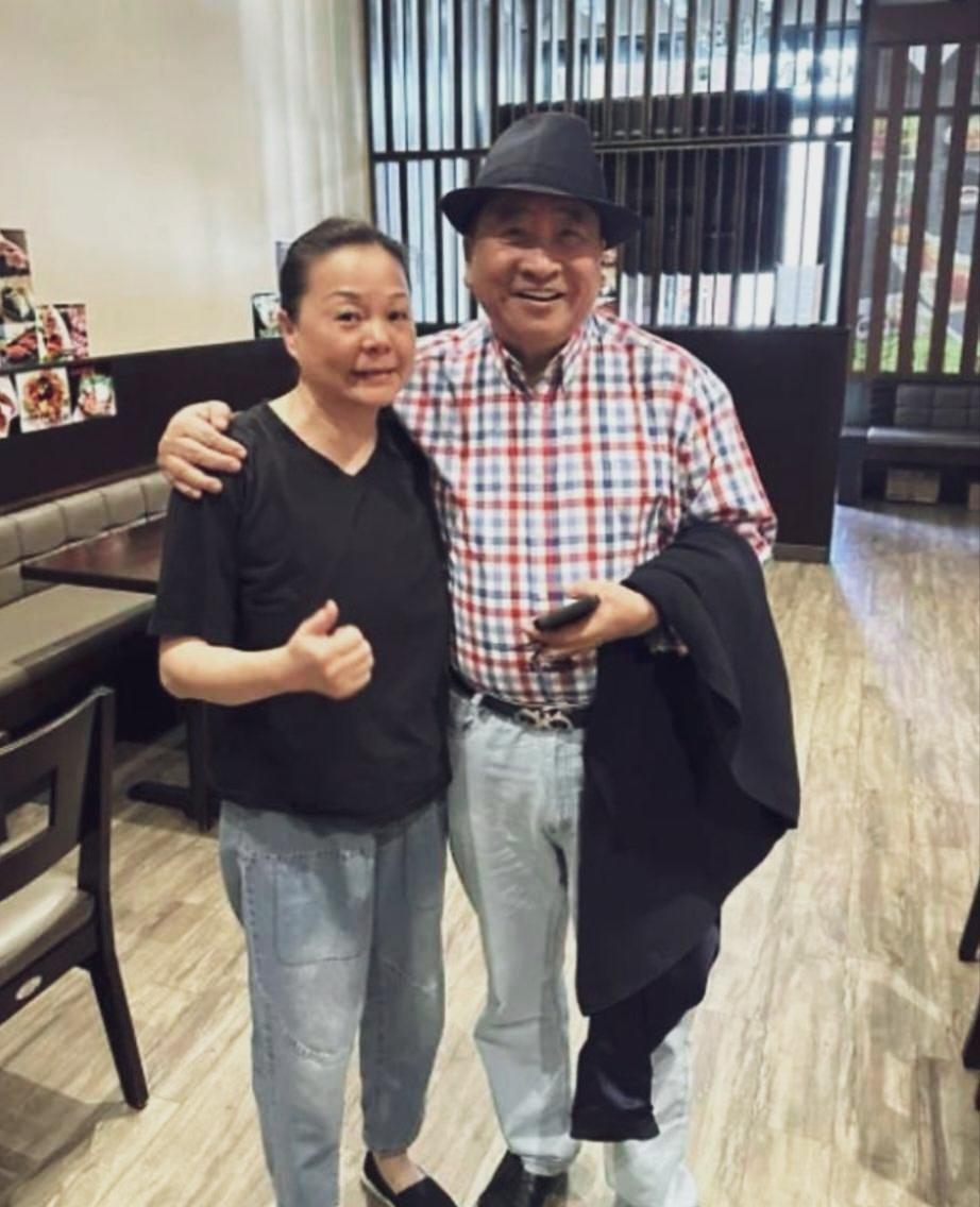 75岁姜昆头戴黑礼帽，身穿红白格子衬衫，身材发福，脸盘子又黑又大，一手搂着身边的