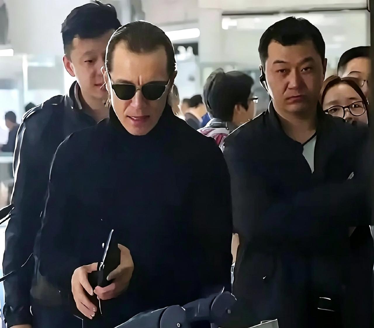 这是李咏出国治病前，在机场拍下的照片。一身黑衣，戴着墨镜，身旁跟着随行人员，谁都