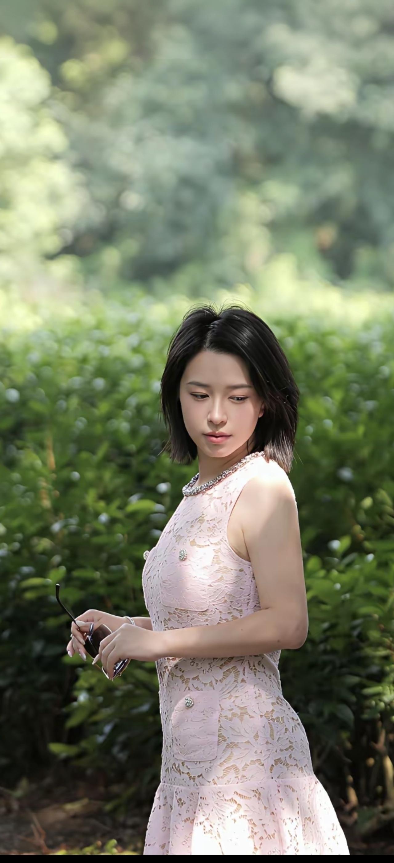 这位女士穿着粉色连衣裙，站在绿植旁。