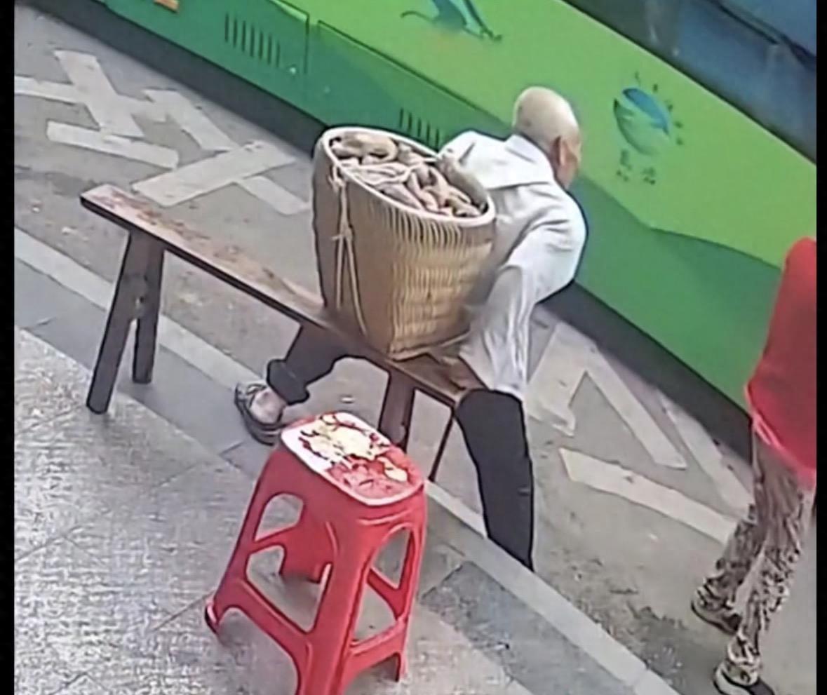 重庆，一老人拄着拐杖、背着竹篓，在超市附近的公交站台苦苦等待，竹篓装满东西有点重