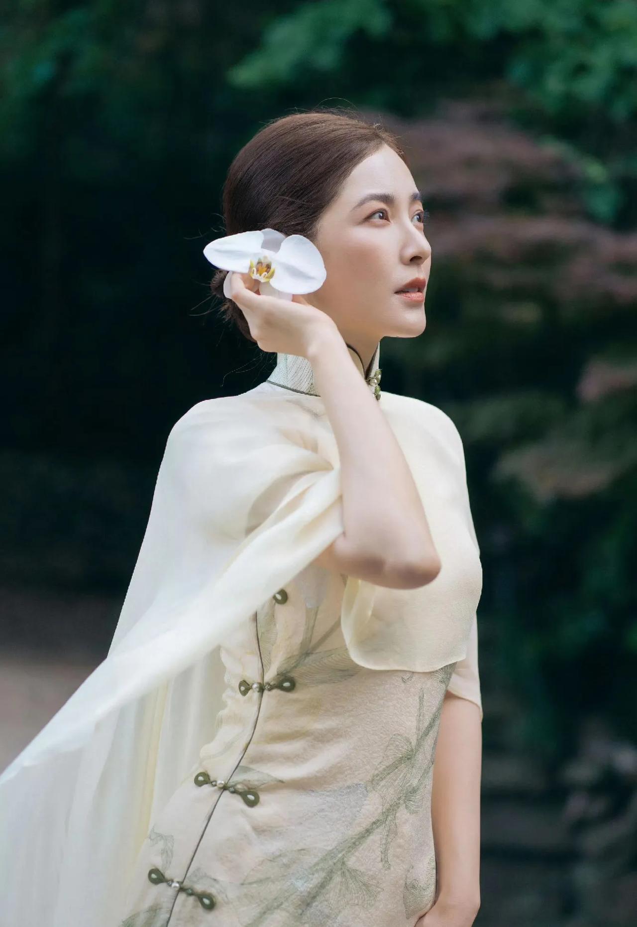曾黎这套浅杏旗袍搭配轻薄外披，携白花置身林间古景，柔和光影衬出温婉古韵，岁月沉淀