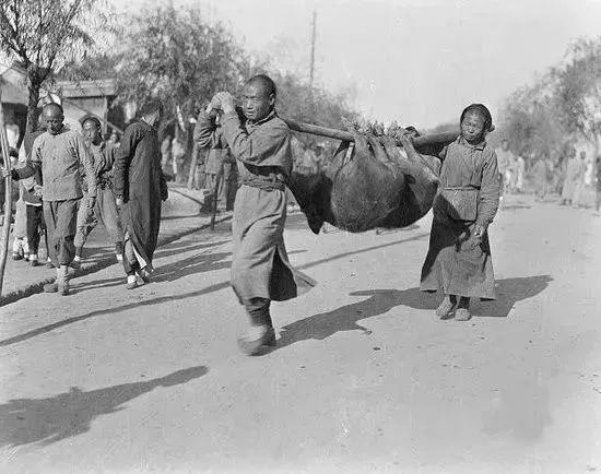 这张黑老照片定格了晚清市井街头的一幕，两名身着粗陋布衣的民众，用长木棍合力抬着两