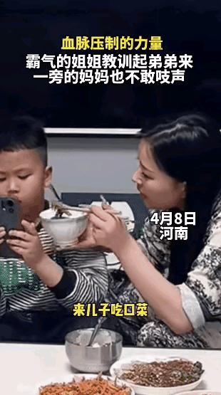 河南，弟弟吃饭时只顾玩手机不愿意吃饭，妈妈端着碗把饭喂到嘴边：来，儿子吃口菜。弟