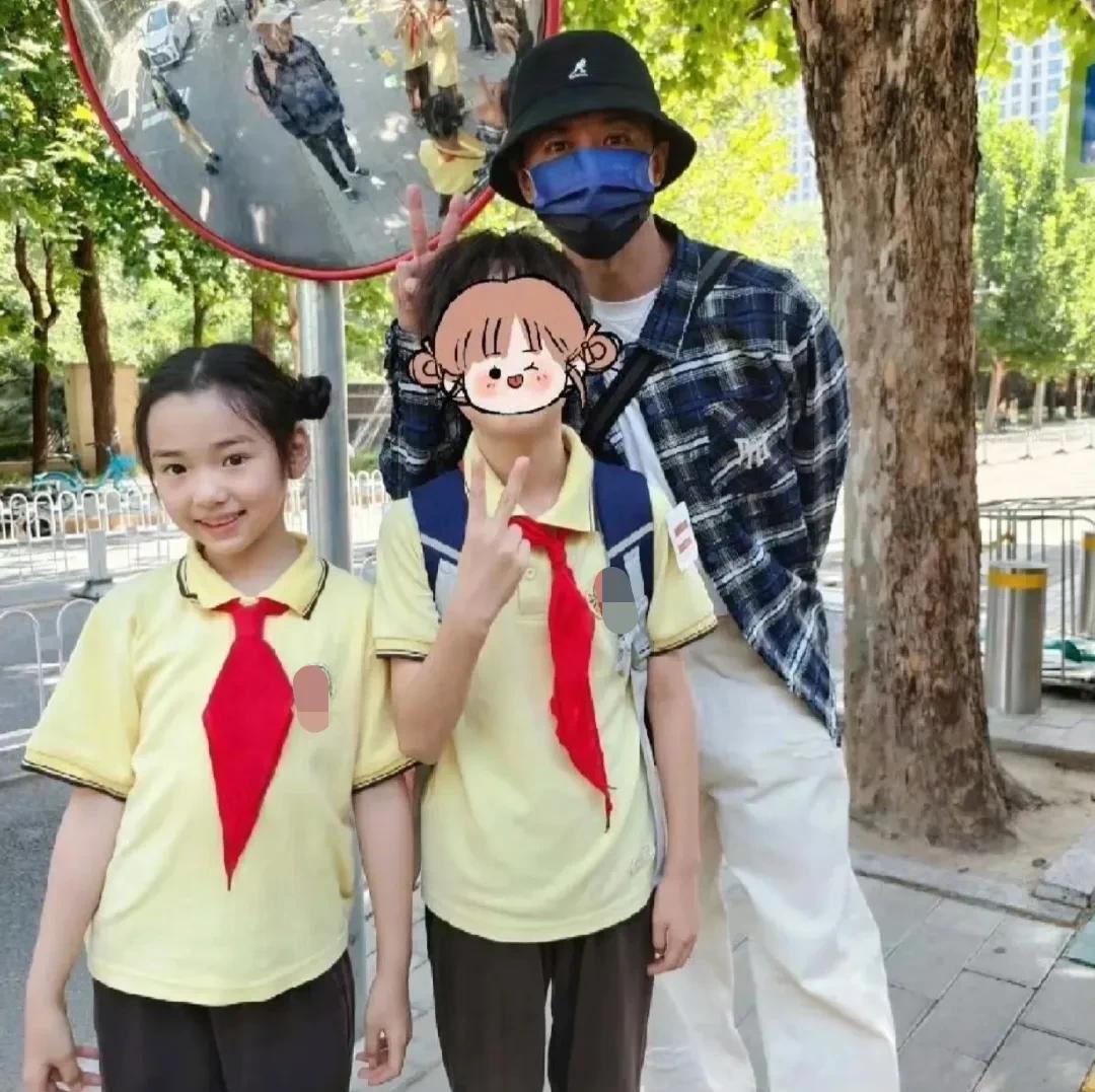 张维伊在北京，接继女小酒窝放学。他戴着渔夫帽和口罩，穿着格子衬衫白色裤子，这穿