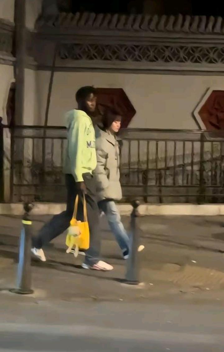 一非洲人和一个女子偎依着在街上走着。看非洲人的表情，很幸福很自豪很满足的样子。