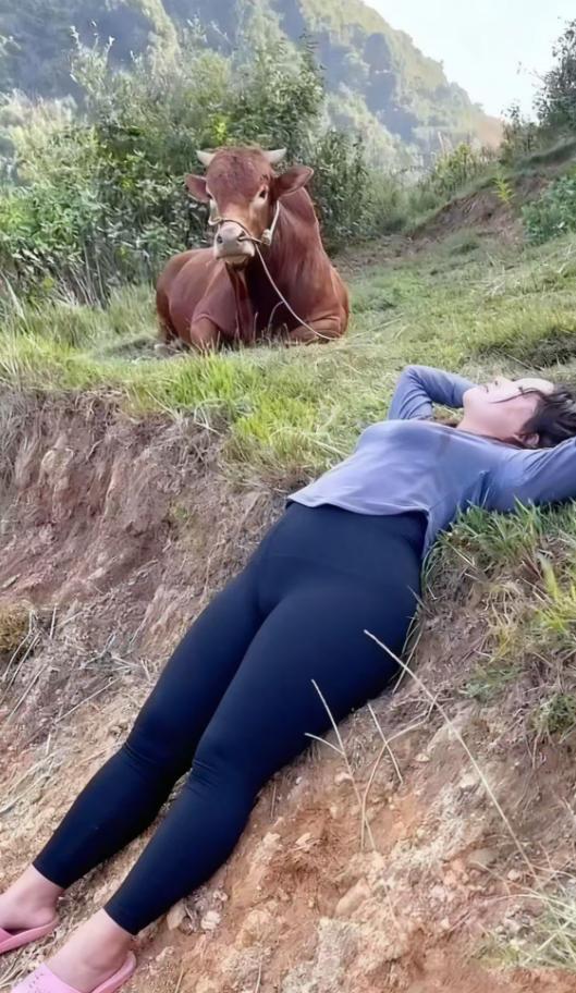 山野坡头，草色混着浅土色铺成软毯，她裹着灰蓝上衣、黑裤往坡上一躺，粉拖鞋松松搭在