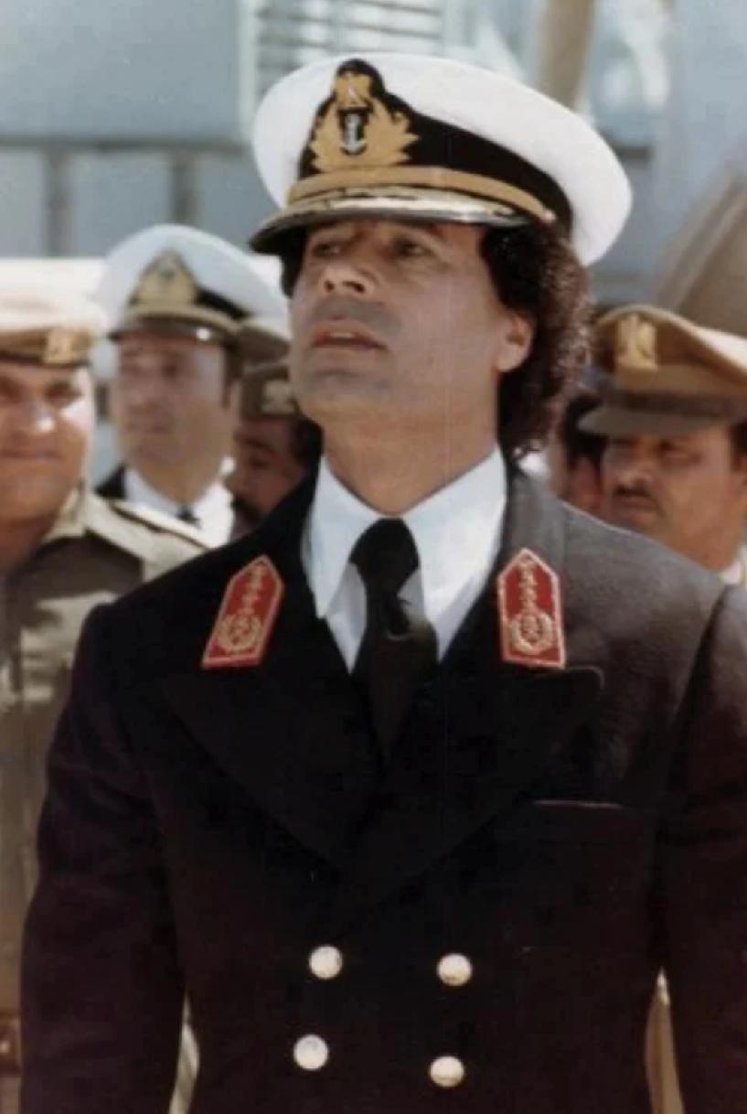 卡扎菲戎装在身，昂首挺胸，意气风发，是个美男子。卡扎菲坐在众多美女中间，穿着休