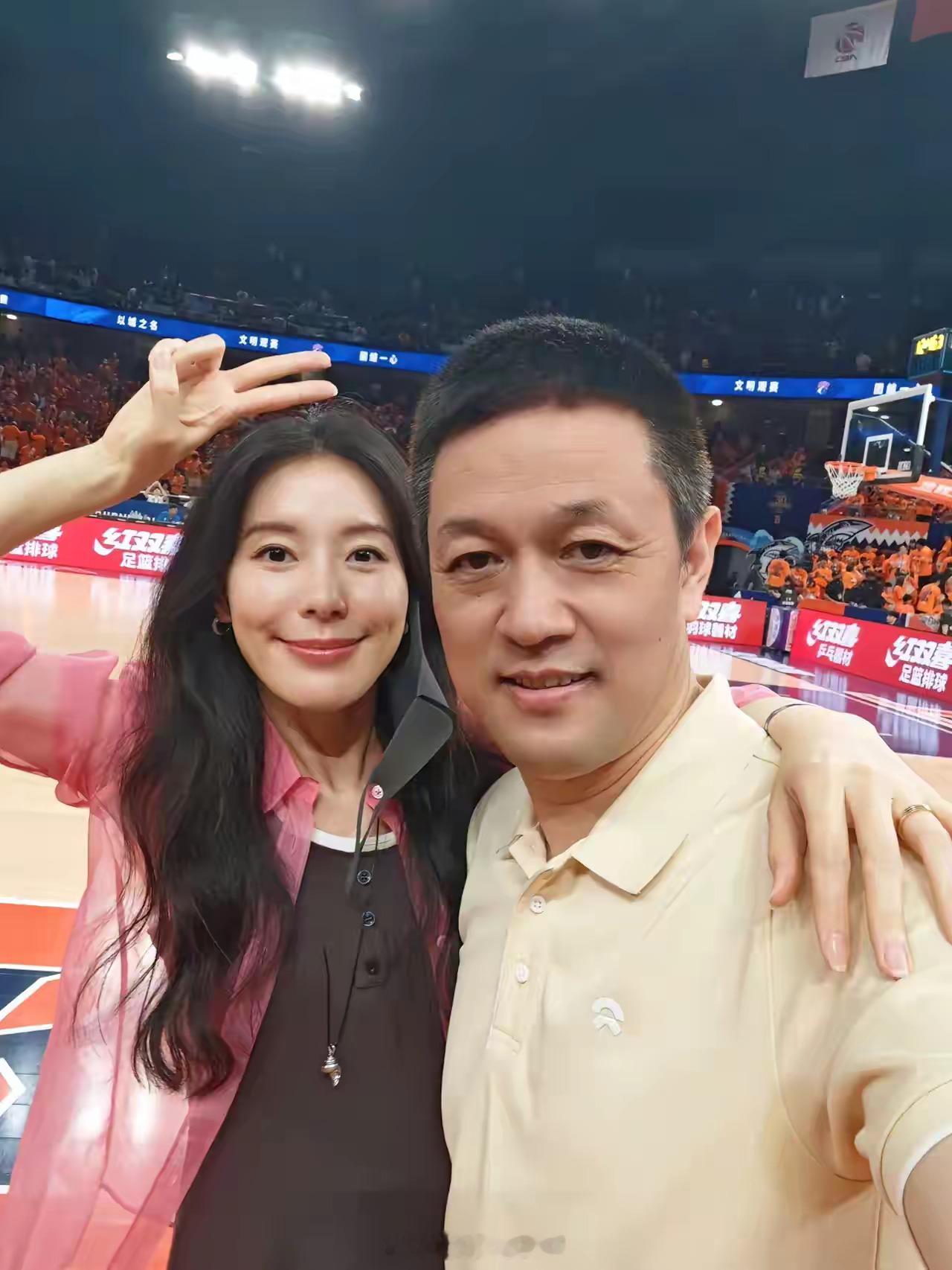 CBA总决赛，观众席上王奕芝身穿粉色衬衫，搭配黑色背心，左手搭在蔚来老板李斌的肩
