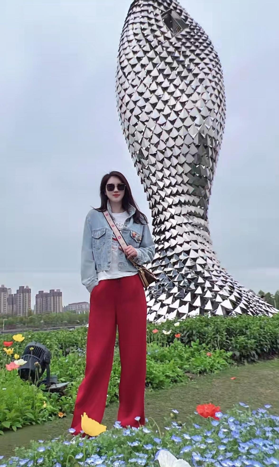 在花丛中，她穿着牛仔外套和红色裤子，站在一个巨大的雕塑旁。