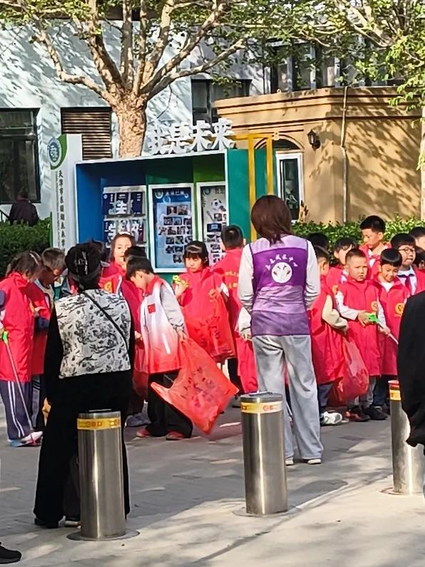 小小志愿者活动在学校门口举行，小同学们在老师的带领下，穿着红色的带有志愿者标志的