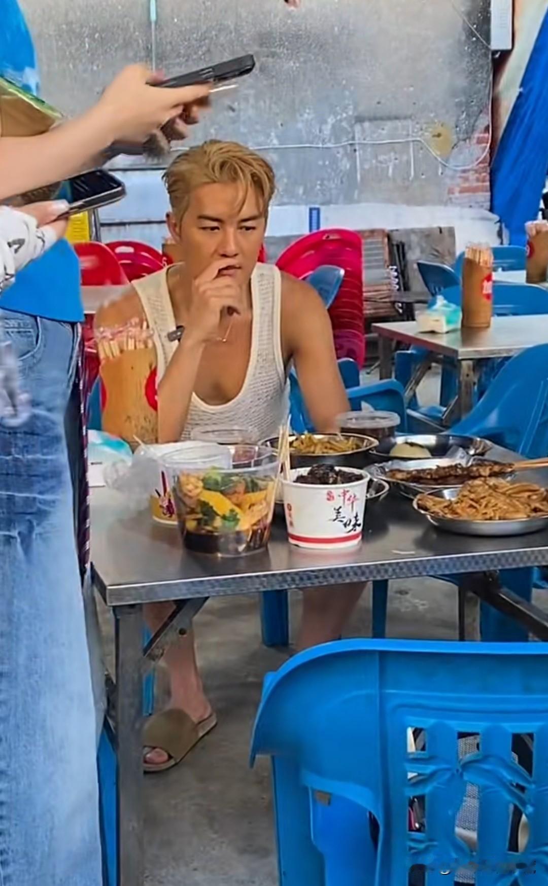 吴克群在大排档吃饭，身穿白色背心，下穿短裤，脚上一双灰色拖鞋的吴克群十分随意，坐