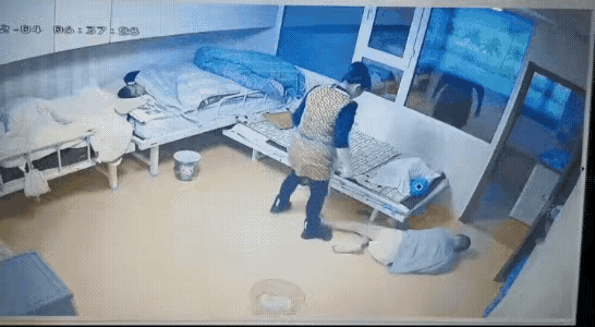 养老院的一个房间，一个老人穿着三角裤躺在地上，一个女护工正在他旁边，用脚踩着抹布