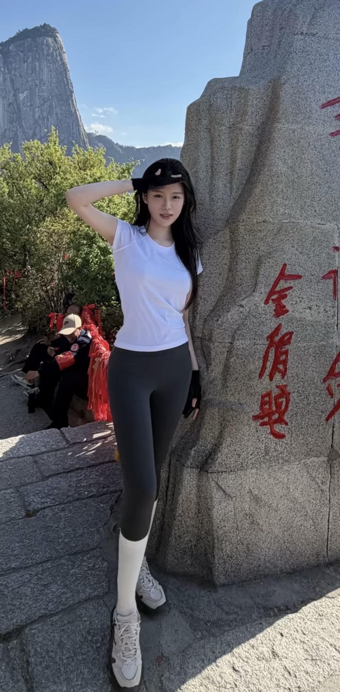 一位年轻女子站在刻有红色书法的巨石旁，背景是苍翠山林与陡峭山峰。她身穿白色短袖T
