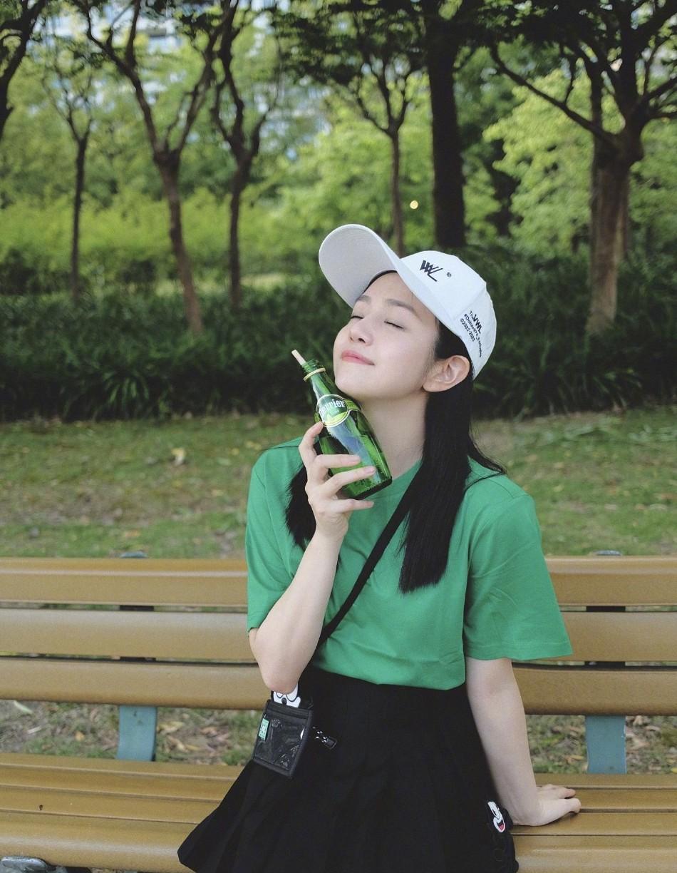 陈妍希发身穿绿色半袖T恤搭黑色半裙，漫步公园喝饮料悠闲惬意。