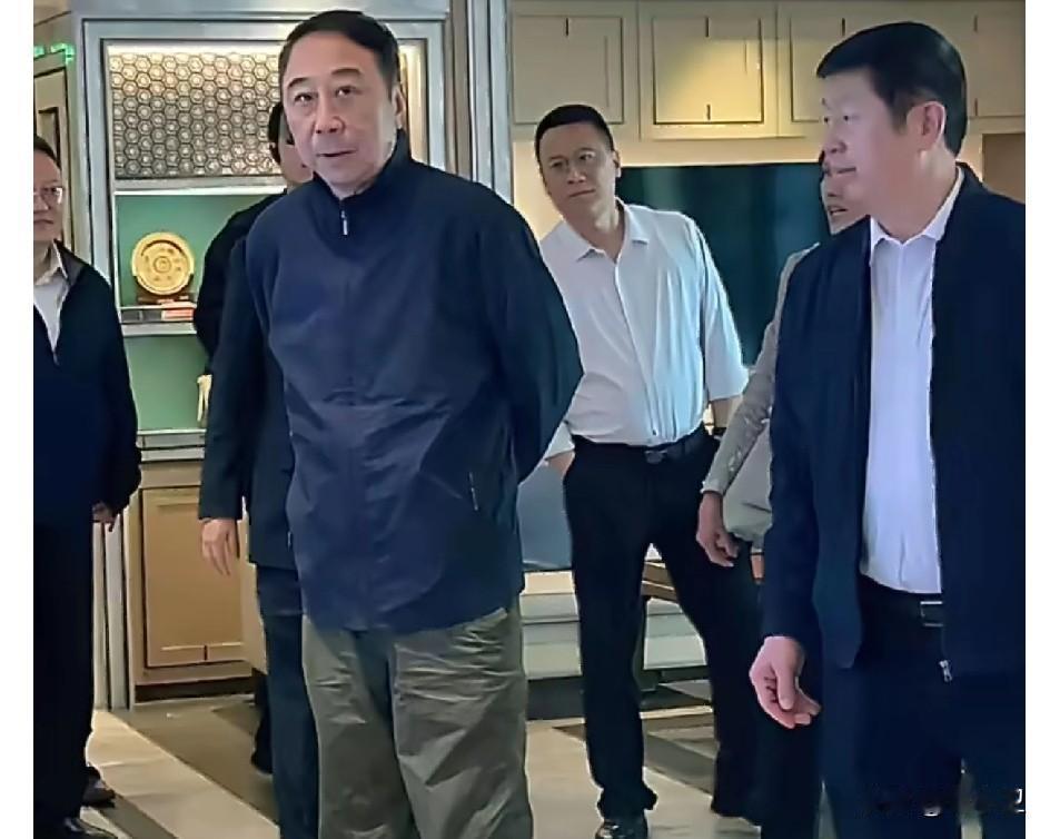 画面里是曲协主席冯巩下基层调研的场景，他身着夹克外套，搭配军绿色长裤，神情严肃庄