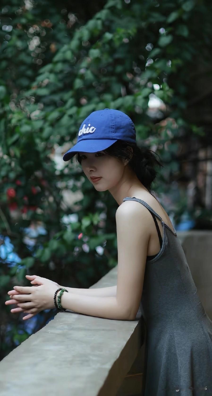 都市少女在绿意盎然中沉思，蓝色棒球帽与灰色连衣裙相得益彰，静谧而优雅。