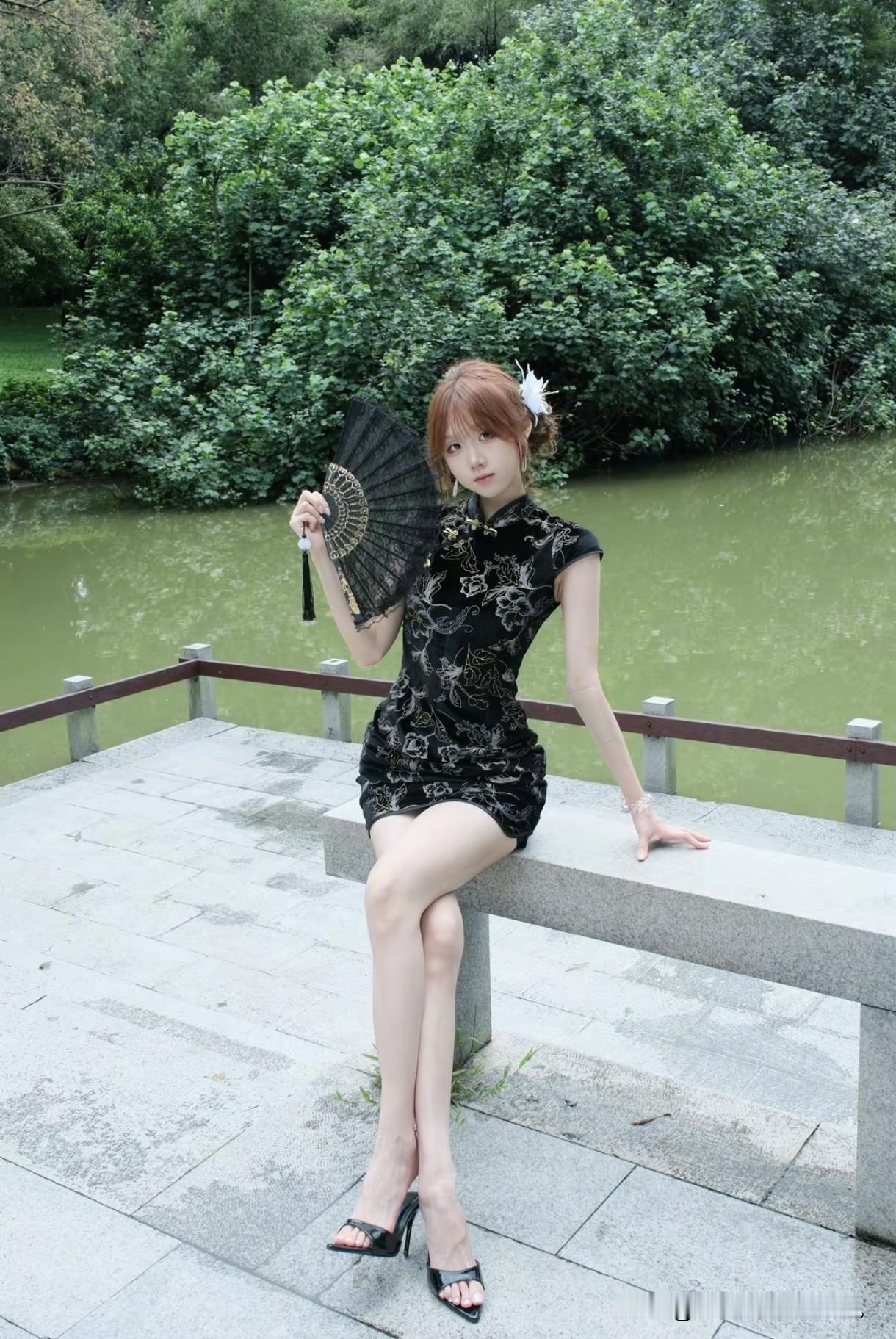 身着黑色旗袍，手持团扇轻摇，坐在湖边的石凳上。绿树成荫，碧波荡漾，这身装扮真是优