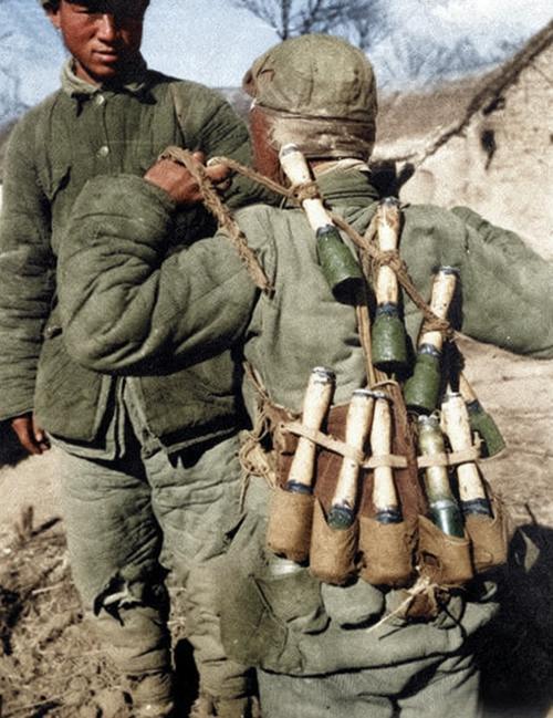 战场土坡上，穿旧棉服的战士背对着镜头，瘦小的肩背被一捆捆手榴弹坠得微塌，木柄磨得