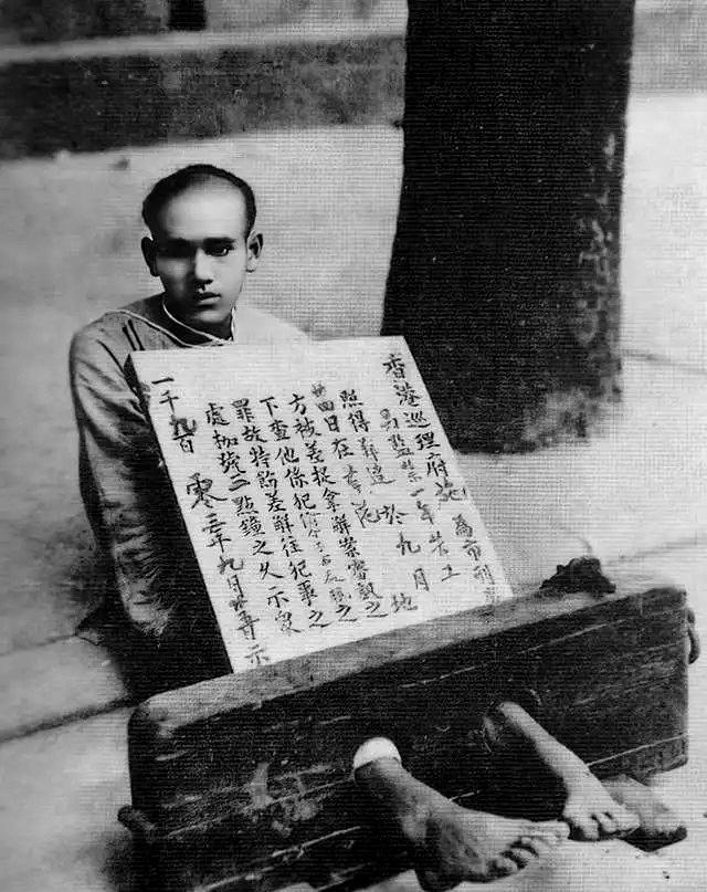 1903年9月25日，香港街头，一名男子坐在路边。他的双脚套着沉重的木枷，脖子上