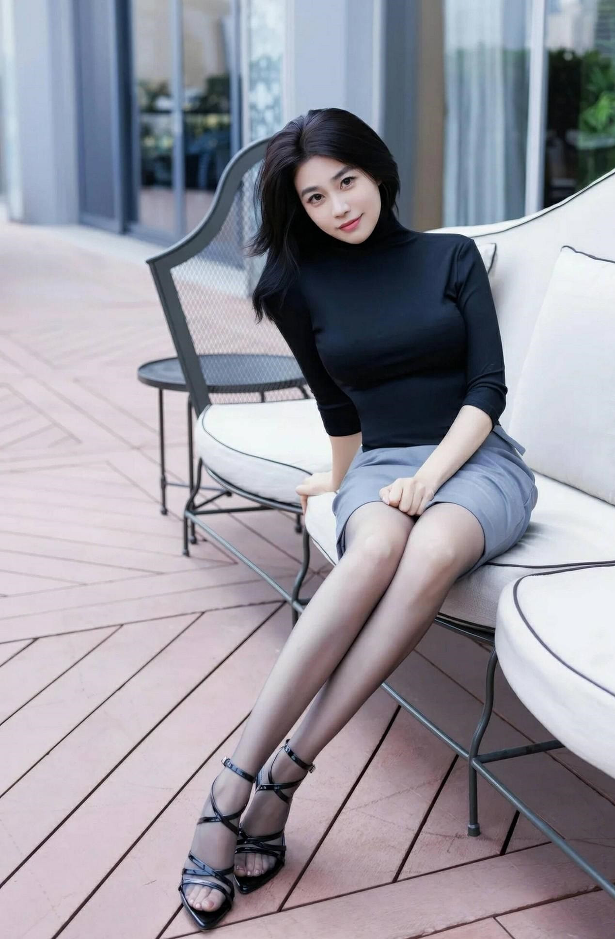 美女身着简约黑高领衫与浅灰开衩裙，优雅坐于户外沙发。黑发轻垂，手抚发丝，黑丝配细