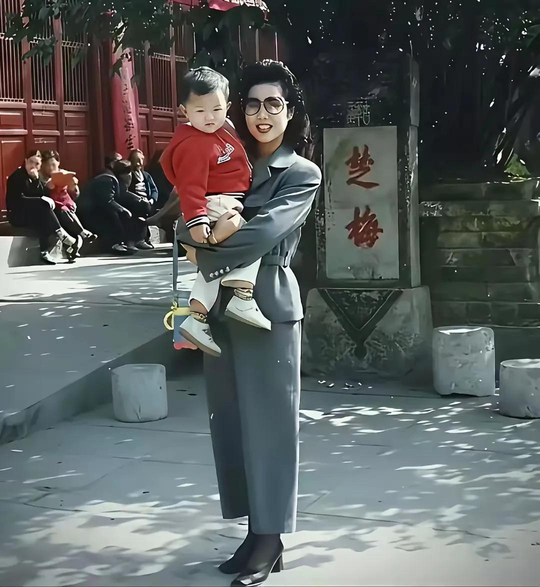 这是当年梦鸽老师抱着儿子在游玩时的留影，穿着打扮上很朴素，手里还拎着个小孩水壶。