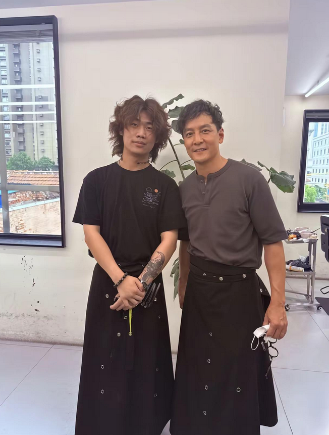 51岁吴彦祖现身上海，身着裙装体验理发师工作，被拍时皮肤略显粗糙。