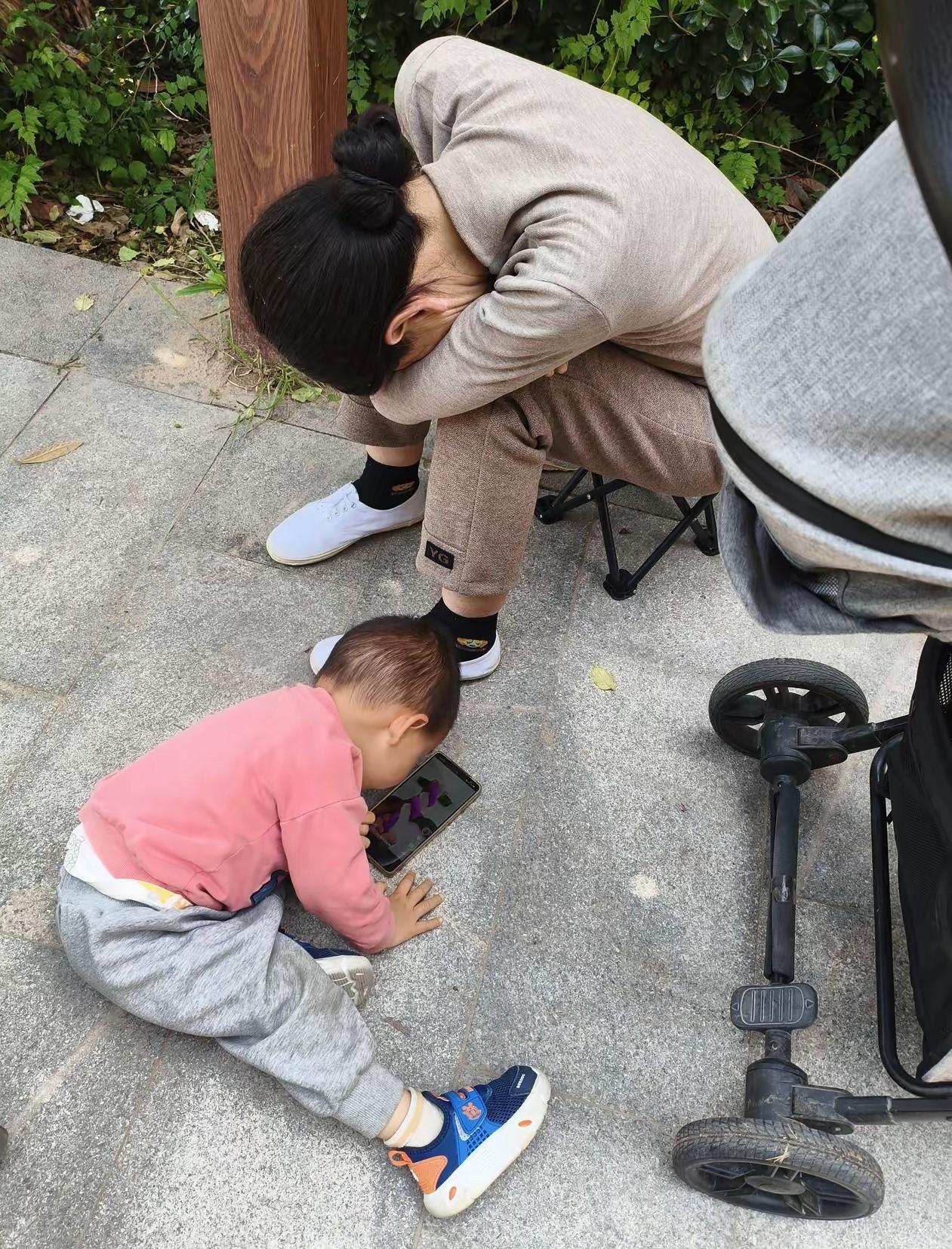 带娃的奶奶好辛苦，奶奶坐在小凳子上睡着了，孩子一个人在玩手机。被网友们拍了下来，