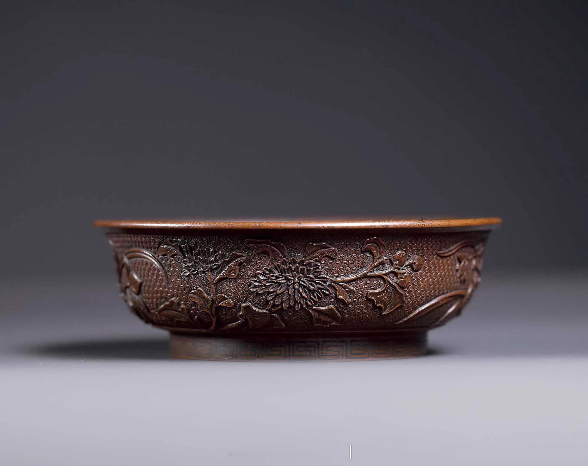 明铜锦地花卉纹碗口沿内侧及足墙以嵌银丝装饰连续回纹，外壁饰锦地浮雕：牡丹、芙蕖
