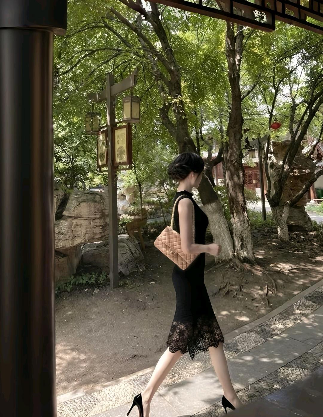 一个美女穿着黑色连衣裙，走在绿树成荫的小路上。