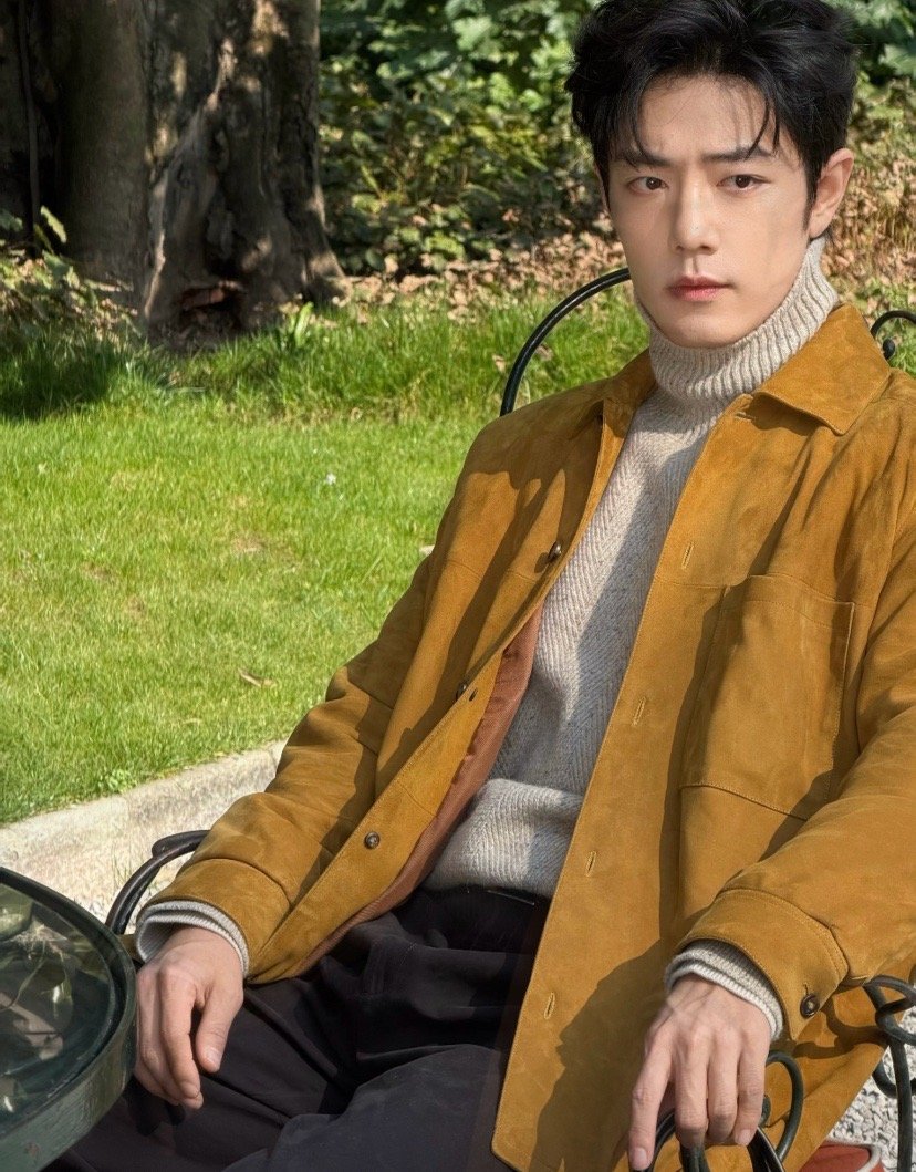焦糖色麂皮外套搭配浅灰高领毛衣，在绿意盎然的庭院里，肖战的松弛感与秋日氛围完美融