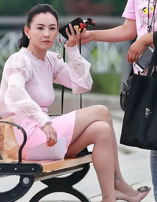 张柏芝穿着粉色短裙脚踩同色高跟鞋扎着马尾辫右手提起手机放到耳朵旁边坐在户外长凳