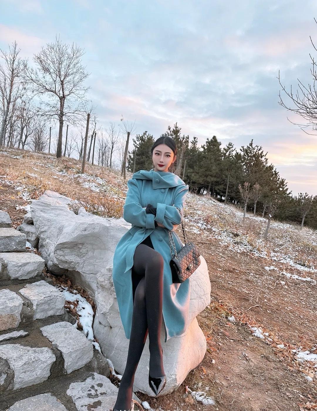 冬日暖阳下，优雅的蓝绿色大衣搭配黑色丝袜，坐在石头上，享受宁静时光。