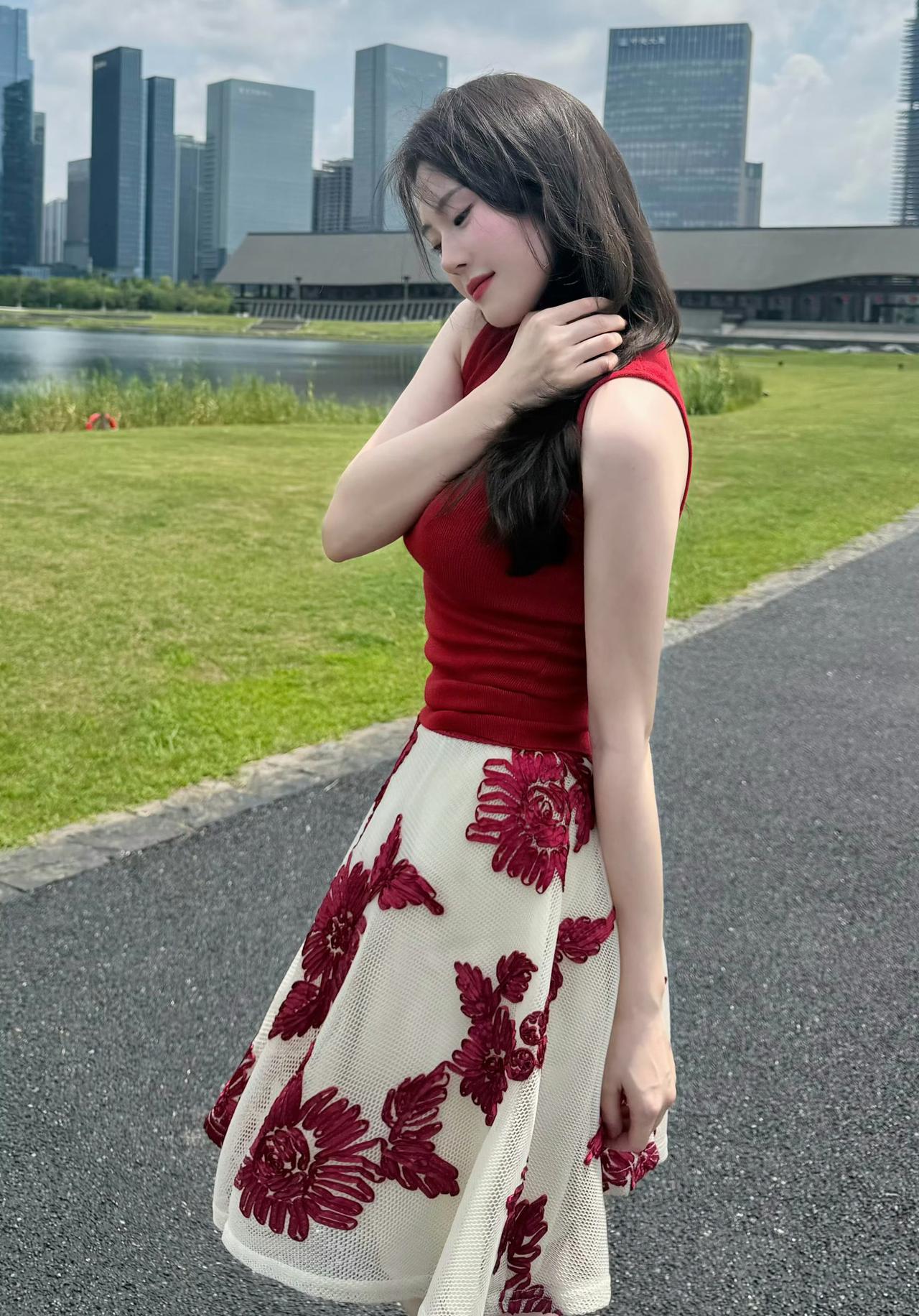 她身着红色无袖上衣搭配带红色花纹的米白色短裙，脚踩黑色夹脚凉鞋，手持闪钻手提包，