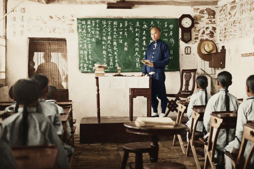 这张老照片拍摄于清末民初一所新式小学堂教室，一位身着长袍马褂的先生站在讲台前，手