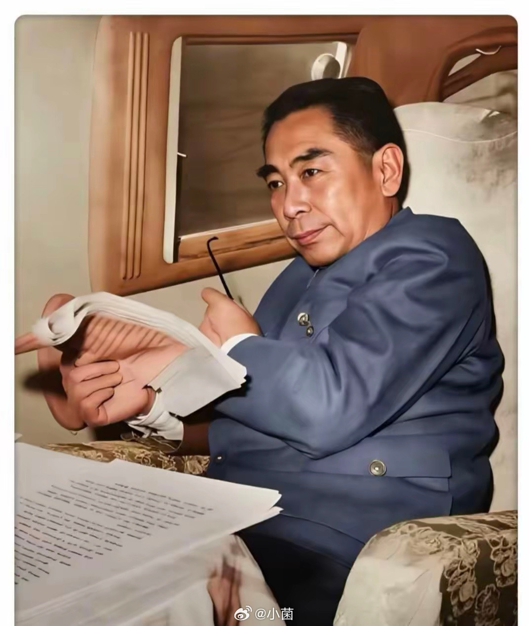 周总理旅途中，身着蓝色中山装，端坐座椅，手持文件与眼镜，凝神阅读。即便在奔波的路