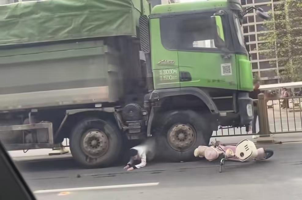 一辆大货车和一辆电动摩托车发生了碰撞，摩托车散架了，女车主被摔到大货车脚去了，周