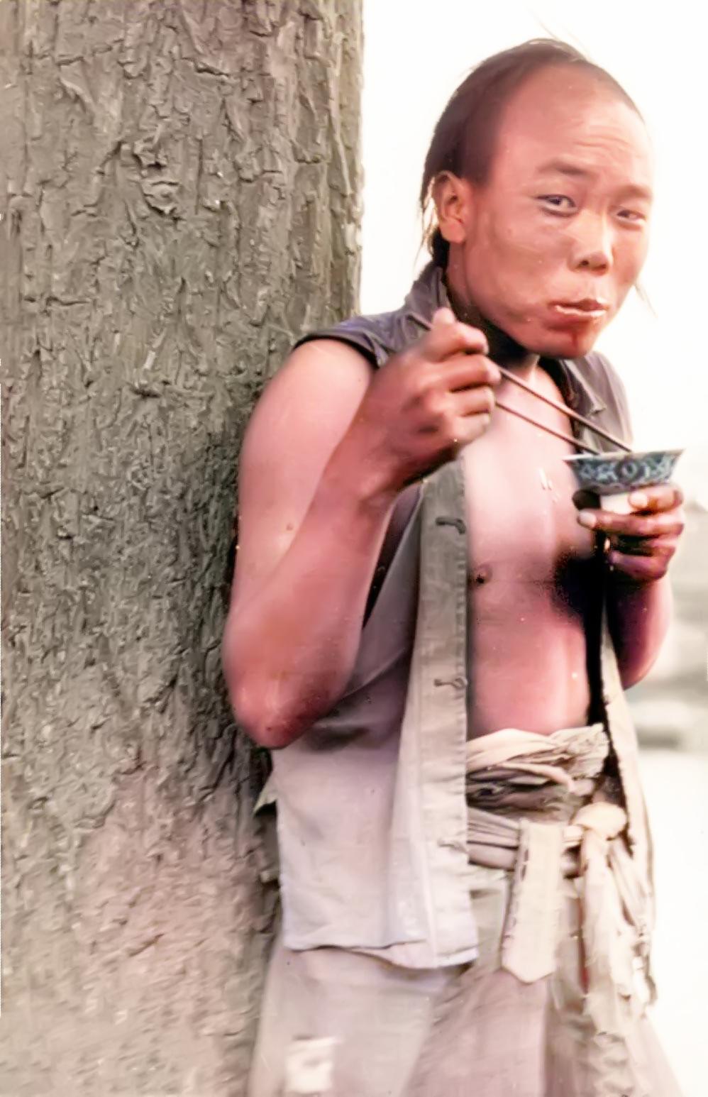 1909年，北京。一个年轻的苦力靠着大树正在吃饭，一手拿着筷子，一手端着装饰有花