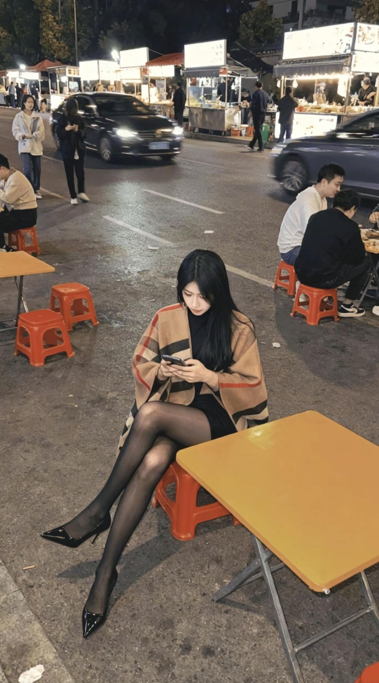 夜色裹着烟火气漫开，路边摊的塑料凳上，裹着格纹披肩的姑娘垂眸划动手机。红凳子、黄